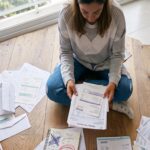 Una mujer sentada en el suelo de madera de un apartamento, revisando facturas y un cuaderno que clasifica la deuda buena vs deuda mala